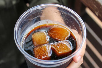 Closeup of hand holding a plastic clear cup with condensation dark black brown coffee cold brew soda drink beverage with four ice cubes
