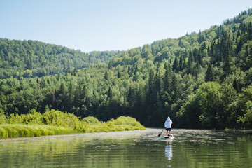 A man is paddleboarding on a serene lake surrounded by lush forested hills, creating a tranquil and idyllic scene of peaceful reflection and outdoor recreation - Powered by Adobe