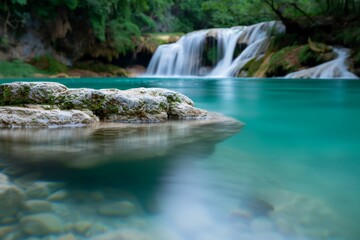 Fototapeta premium Scenic view of a waterfall cascading into a serene turquoise pool surrounded by lush greenery and rocky formations.