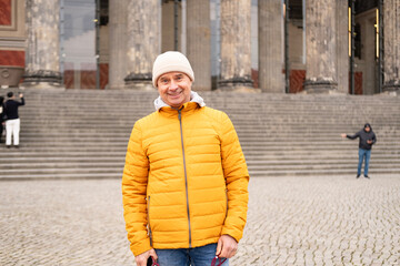 Cheerful middle-aged man, 65 in winter bright yellow puffer jacket and beanie, smiling on urban...