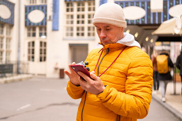 Cheerful middle-aged man, 65 in winter bright yellow puffer jacket and beanie, smiling on urban street with historic buildings in background © kittyfly