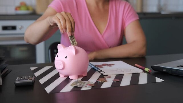 Young woman couting coins and banknotes while sitting at desk in the kitchen, putting it into a pink piggy bank. Savings and budgeting concept