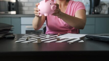 Young woman empties a pink piggy bank, scattering coins onto a table covered with papers and a laptop. Concept of budgeting and personal finances