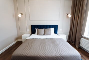 In the center of the room, there is a bed with a blue headboard. It enhances the comfort and style of the space, surrounded by other furniture and fixtures like curtains and lighting