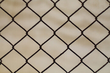 Fototapeta premium Dark side of metal mesh fence, behind blurred beige background. Edges of contours create silhouette, symbolize closed borders, without breakthrough of it will not be possible to see further