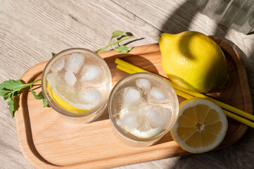 Summer refreshing lemonade drink with ice, lemons on wooden tray in bright natural sunlight with shadows, top view, lifestyle food composition