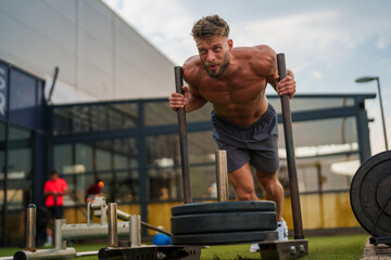 Muscular caucasian fitness instructor pushing a weighted sled during an outdoor workout at a gym.