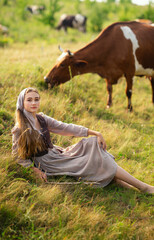 Beautiful blonde girl in scarf and  medieval rural clothes on a pasture with a cows. Farm life.Young model posing on a ranch.Warm art work.