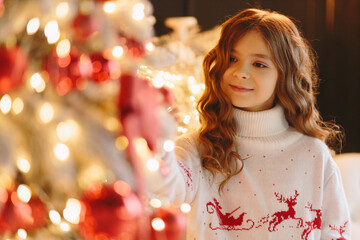 A cute girl in a white sweater with deer looks at the camera against the background of a Christmas tree.