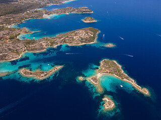 Aerial view of an island on Sithonia Peninsula, Greece, with beaches and rocks, surrounded by crystal-clear waters. Yachts and boats anchor in turquoise waters. Nearby islets and reefs visible