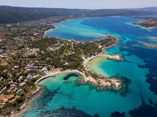 Aerial view of an island on Sithonia Peninsula, Greece, with beaches and rocks, surrounded by crystal-clear waters. Yachts and boats anchor in turquoise waters. Nearby islets and reefs visible