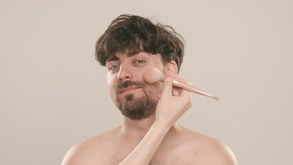 Young adult man looks at camera female applies powder on his face., isolated on white background. Natural beauty skin care concept.