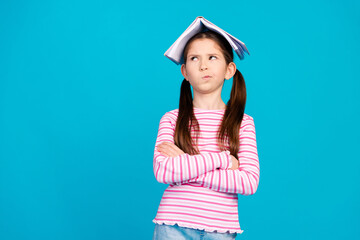 Photo of minded small girl dressed striped shirt holding book on head arms crossed look empty space isolated on blue color background