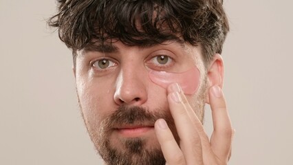 Young adult man applying eyepatch under eyes looking at camera, isolated on white background. Natural beauty skin care concept.