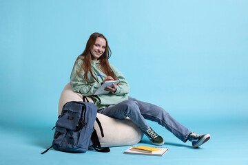 Portrait of radiant teenager sitting on beanbag and writing on notebook, studio backdrop. Upbeat...