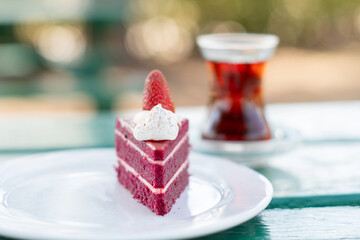 piece of red velvet cake decorated with fresh berries in a white plate on the table, a cup of Turkish tea in a traditional Armudu glass, time for a sweet dessert during the summer holidays