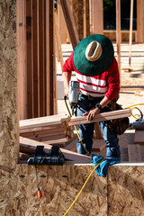 Construction worker in toolbelt and sun hat working with a pneumatic nail gun on new house wood construction framing, summer construction season
