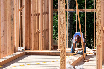 Construction worker in toolbelt and hard hat working with a pneumatic nail gun on new house construction wood framing, summer construction season
