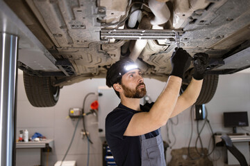 Dedicated mechanic performing maintenance on car in professional garage. Wearing headlamp and gloves, focusing on vehicle's undercarriage for repair work.
