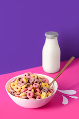 Bowl with colorful sweet cereal rings and bottle of milk on pink table