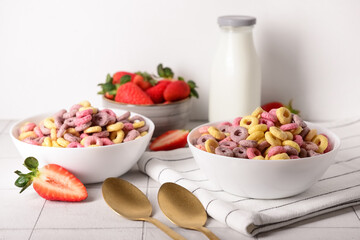 Bowls with colorful sweet cereal rings, strawberries and bottle of milk on white tile table