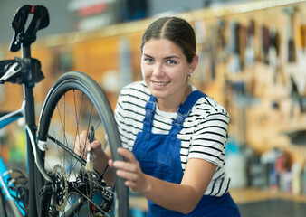 Smiling skilled young female mechanic in blue overalls adjusting bicycle wheel, cleaning gears with small brush in professional bike repair shop