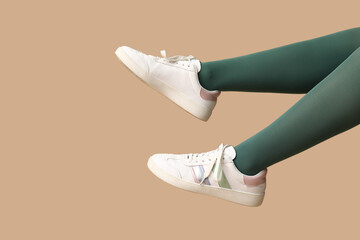 Legs of young woman in green tights and white sneakers on beige background