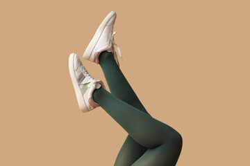 Legs of young woman in green tights and white sneakers on beige background
