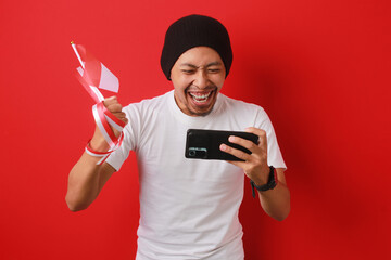 Overjoyed Indonesian man celebrates watching the Indonesia National team score a goal and win the game on his phone while holding the Indonesian flag during Indonesia Independence Day on August 17