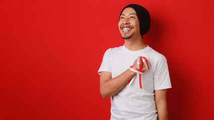 Happy Indonesian man in a white T-shirt and beanie hat, with an Indonesia flag ribbon, puts his...
