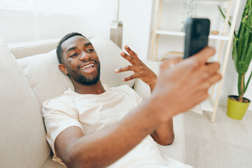 Happy African American Man Sitting on a Black Sofa and Using a Mobile Phone in His Modern Apartment He is relaxed and smiling while typing a message, enjoying the comfort and connection provided by