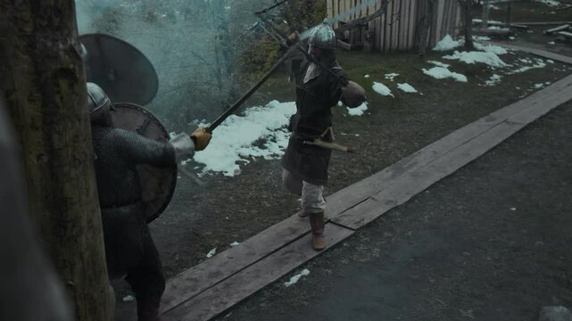 Full high-angle slowmo shot of medieval male warrior in helmet, with two swords fighting with two enemy intruders wielding shields and axes, in clouds of smoke, during ambush on fortress