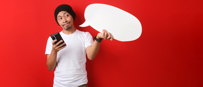 Thoughtful Indonesian man ponders while looking at a speech bubble in his hand, considering the words to type on his phone to update his status about Indonesia Independence Day on August 17