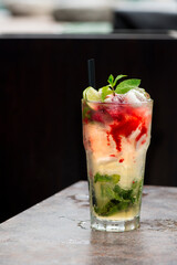 A glass of mojito with a red straw rests on a wooden table. The glass is filled with light green liquid, mint leaves, and raspberries.
