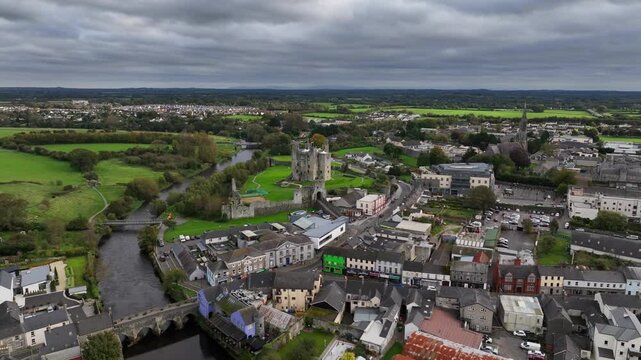Trim Castle, County Meath, Ireland, October 2023. Drone orbits clockwise in a wide panoramic aerial establishing the Norman Stronghold located in a bustling town on a cloudy day.