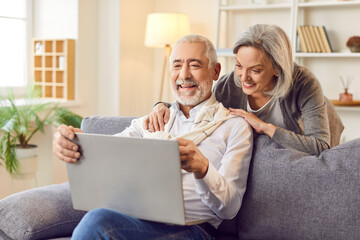 Elderly couple is spending leisure time together, using a laptop and laughing, sitting on a sofa in the living room at home. They are engaged in online entertainment, enjoying internet activities.