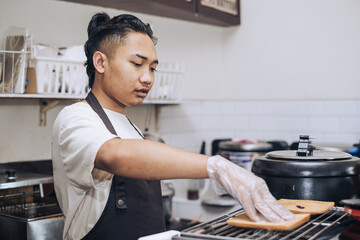 A Southeast Asian chef wearing an apron is toasting two pieces of loaves in the kitchen with bokeh background