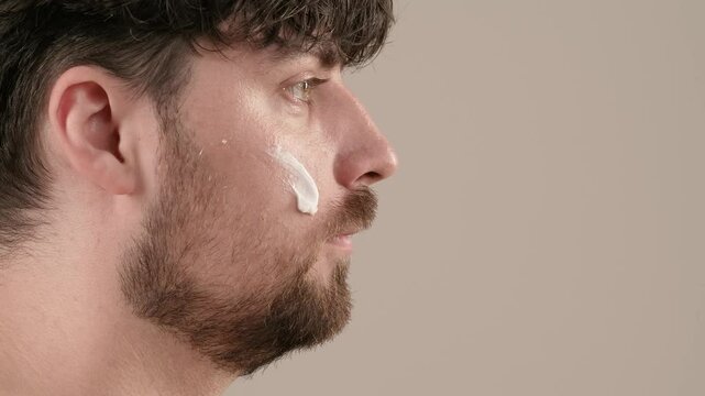 Young adult man applies aftershave cream moisturizer looks at camera, isolated on white background. Natural beauty skin care concept. Side view.