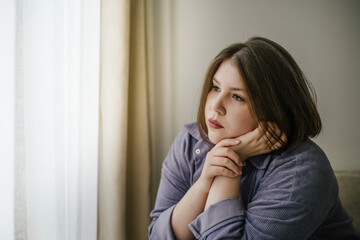 Sad plus-size woman with pensive expression looking out window with sheer curtains. 