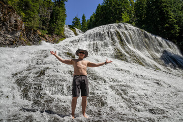 Adventurous athletic man standing in a large waterfall with his shirt off on a bright sunny summer day.