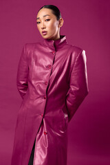 Fashionable asian brunette in purple leather coat posing against purple background with hands on hips