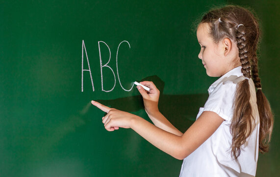 A schoolgirl writes the letters A B C on the blackboard. The girl writes the English alphabet with chalk on a green school board. A child learns to write letters at school. Back to school.
