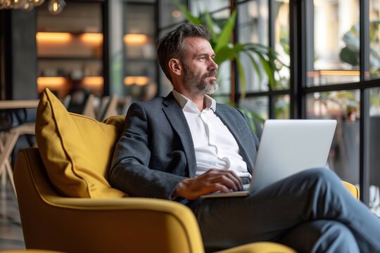 thoughtful businessman looking away while sitting with laptop on comfortable seat at flexible workplace.