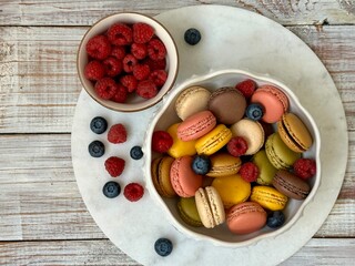 Macarons in different colors and tastes in the white plate and raspberries and blueberries on the marble plate on the wooden background 