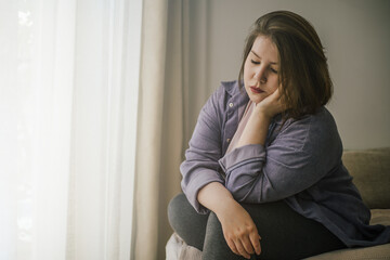 Plus size woman sitting alone by window, wearing casual clothes.