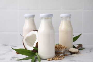 Different types of vegan milk in bottles with ingredients and green leaves on white marble table