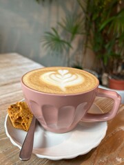 Pretty pastel pink cup of cafe latte with heart latte art on a wood grain table. Placed on a white saucer with a piece of cookie and a teaspoon.