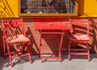 A red table and two chairs are set up outside a brick building, with the chairs facing each other. Red patio set outside brick building.