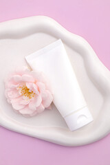 White plastic tube with with a collagen cream on white tray on pink background with blooming rose flower. Natural cosmetics concept, natural essential skin care products