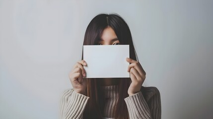 Naklejka premium A young girl holding a blank white card in front of her face, on a light background, concept of anonymity